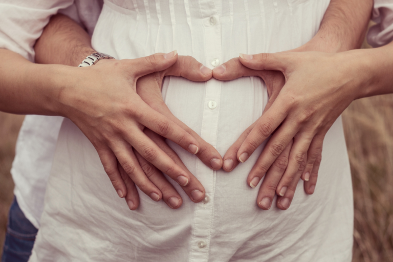 A man's and women's arms round her pregnant stomach, making a heart with their hands 