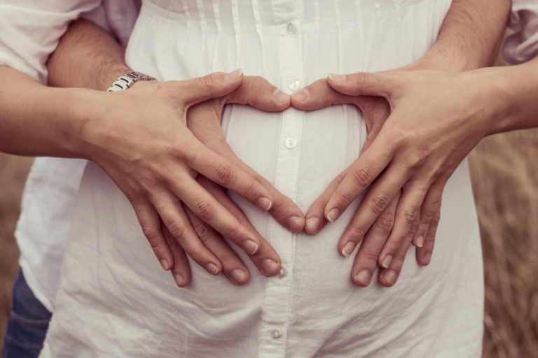 A man's and women's arms round her pregnant stomach, making a heart with their hands