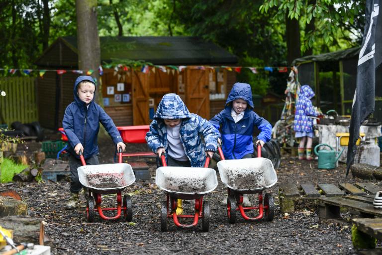 Three children playing outside, pushing wheelbarrows