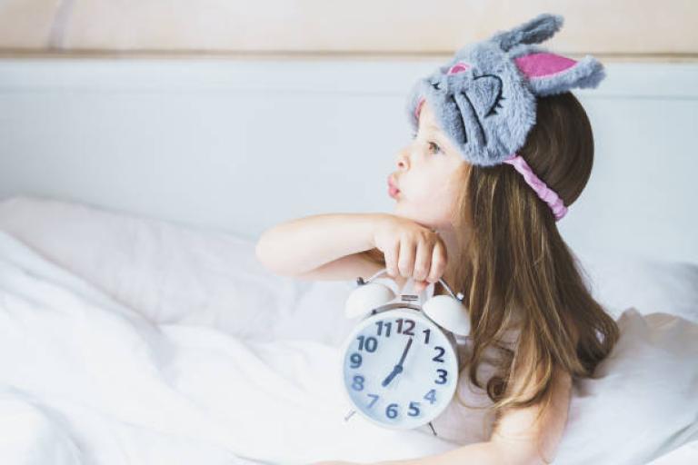 Young child holding an alarm clock, sat in bed with an eye mask on her head
