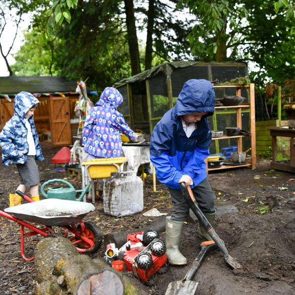 Children play in rain coats and wellies by digging in the soil in an outdoor classroom.